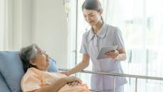 A nurse holding a digital tablet is assisting a patient lying in bed